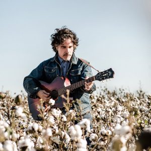 White man in denim jacket playing an acoustic guitar in cotton field