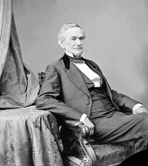 Older white man in suit high collar and bow tie posing in a chair with his elbow on a table covered in ornate cloth