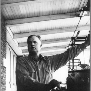White man in button-down shirt standing on covered porch with machinery