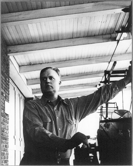 White man in button-down shirt standing on covered porch with machinery