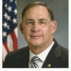 White man with glasses in suit and tie with flag behind him