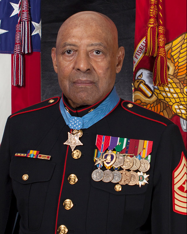 Older African-American man in military uniform featuring medals with flags behind him