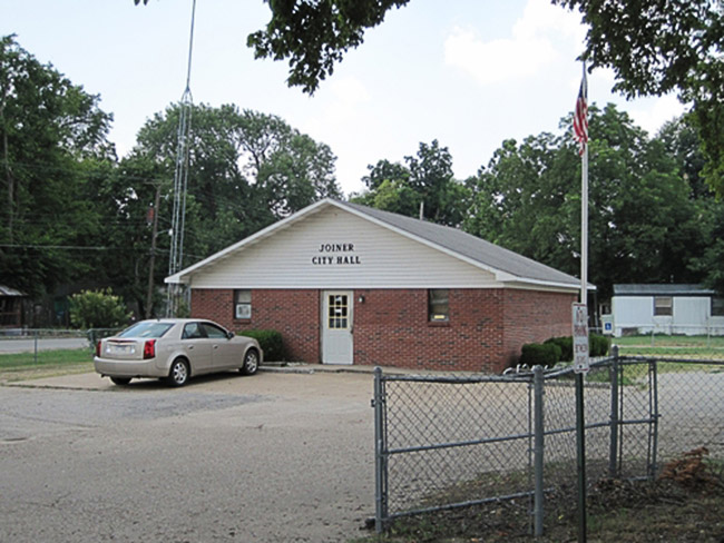 Brick building labeled "Joiner City Hall" with car in parking lot