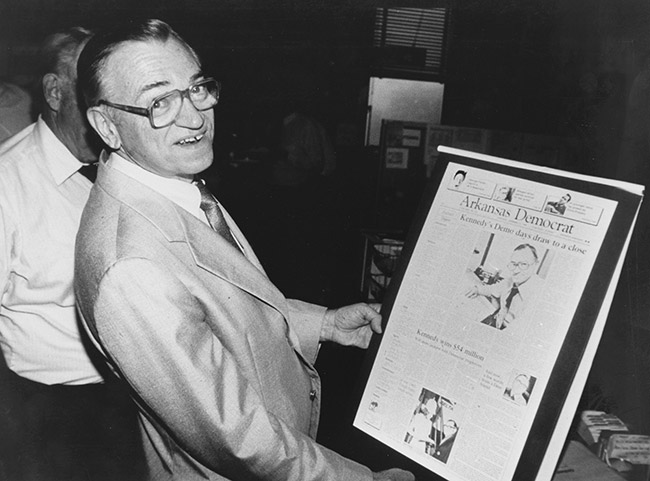 Older white man with glasses in suit smiling as he holds a page from the Arkansas Democrat about him in his hands