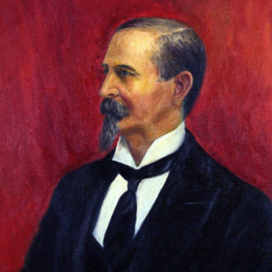 Profile view of white man with mustache and long chin beard in suit before a red background