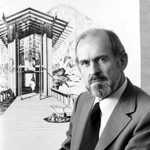 White man with a beard and mustache in suit and tie standing before the drawing of a building