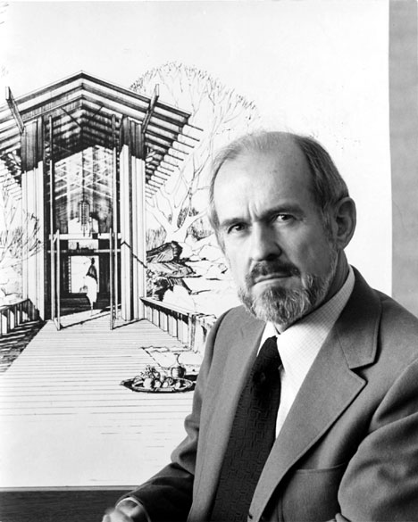 White man with a beard and mustache in suit and tie standing before the drawing of a building