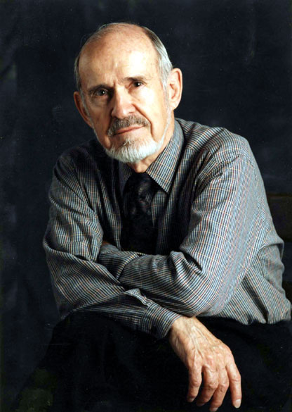 Old white man with mustache and beard in striped shirt and tie with arms crossed in front of him.