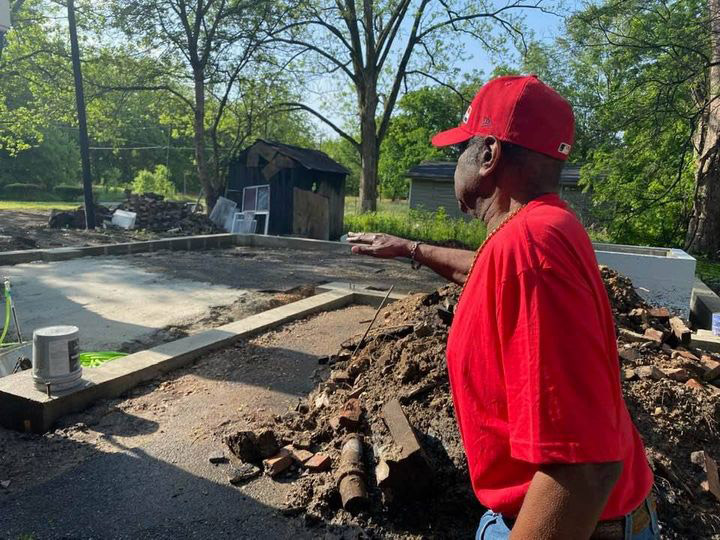 African-American man in red shirt and hat standing next to pile of rubble pointing towards new foundations under construction