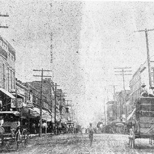 Horse drawn wagons and man on town street with brick storefronts and power lines
