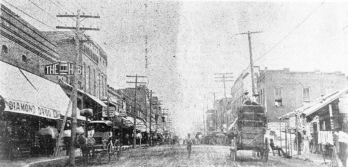 Horse drawn wagons and man on town street with brick storefronts and power lines