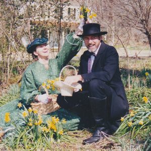 White woman and white man sitting amidst flowers with multistory house in background