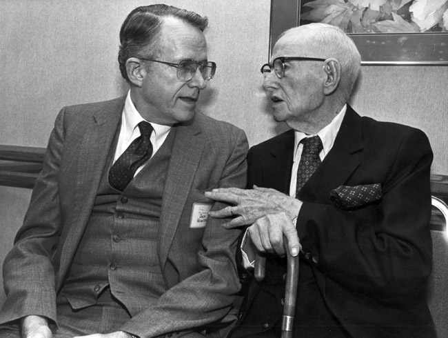 Two older white men in suits and glasses seated conversing with the elder gesturing holding cane