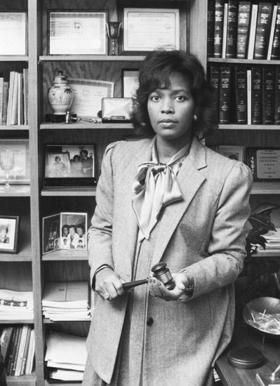 black woman in suit and silk necktie with gavel standing in front of bookshelf