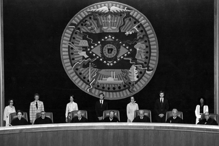 five judges in black robes at the bench with their clerks behind them under State Seal
