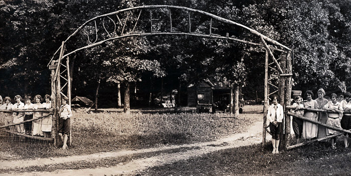 White children at both sides of camp entrance gate with arch above on dirt road