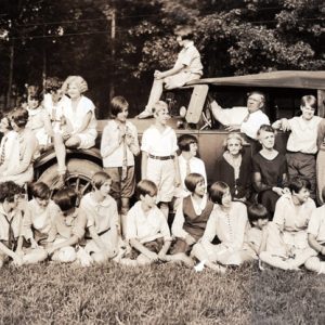 children and adults mostly dressed in white gathered around a car with an older man behind the wheel