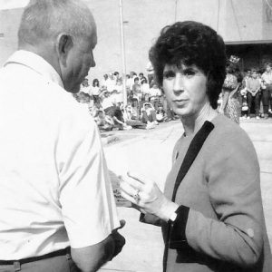 White woman with short hair talking to old white man with crowd in the background