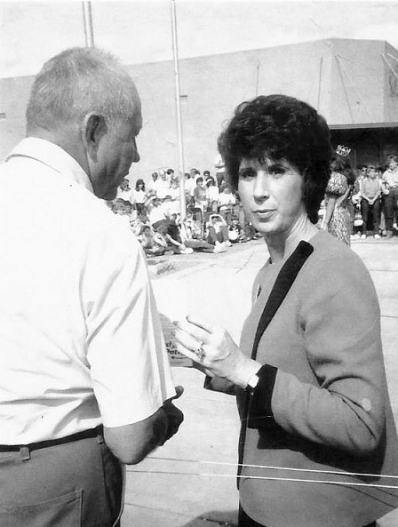 White woman with short hair talking to old white man with crowd in the background