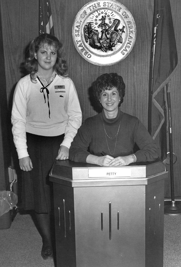 White woman standing next to white woman sitting at desk with State Seal on the wall behind them