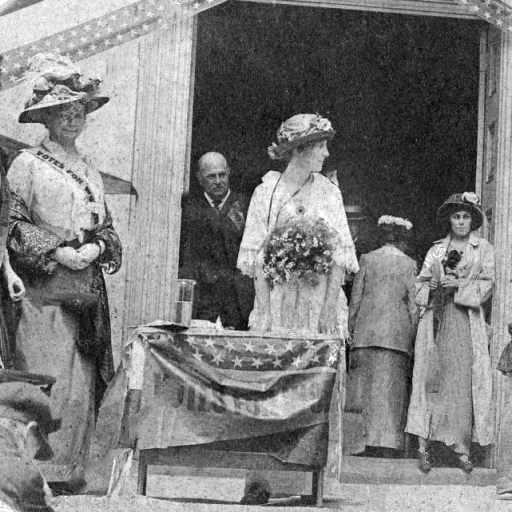 White women in dresses with hats and white man on building steps with flag banner