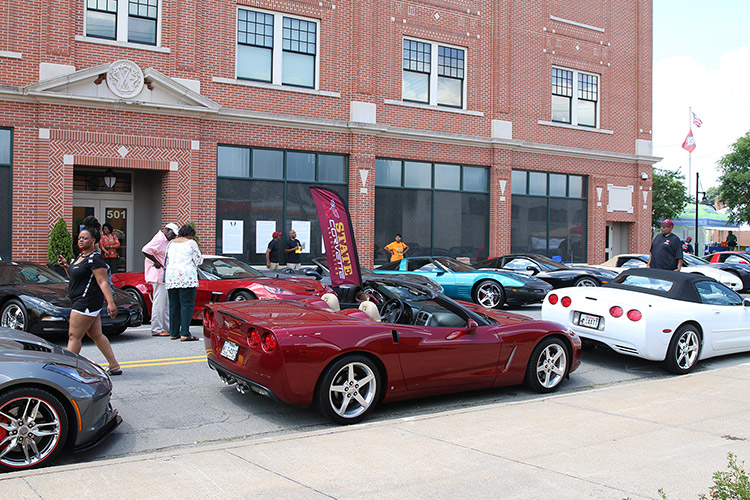 African-American men and women looking at Corvette sports cars on street in front of multistory brick building