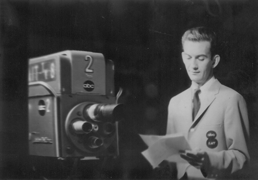 White man in suit and tie reading papers next to television camera