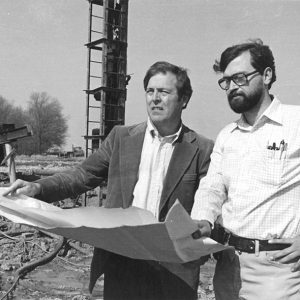 Two white men outdoors looking over large piece of paper with tower behind them