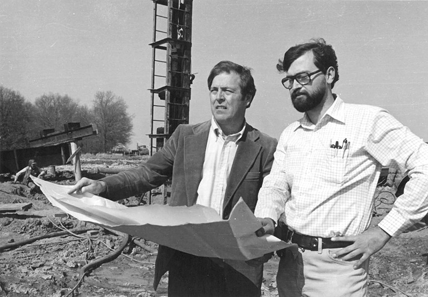 Two white men outdoors looking over large piece of paper with tower behind them