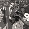 White man in suit and tie holding microphone alongside African-American man behind TV camera