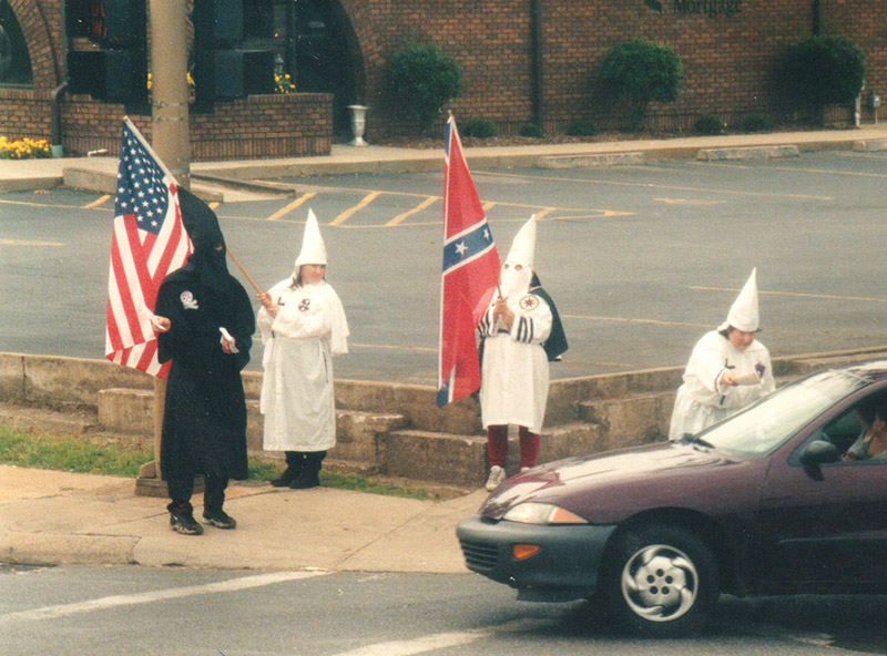 White men wearing Ku Klux Klan outfits and carrying confederate and American flags on street corner