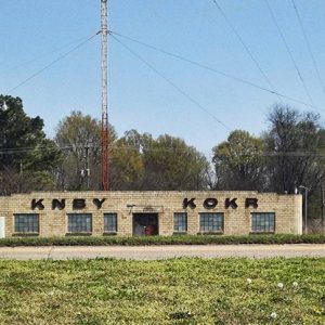 Single-story brick building with letters saying K.N.B.Y. and K.O.K.R. on it on street with radio tower behind it