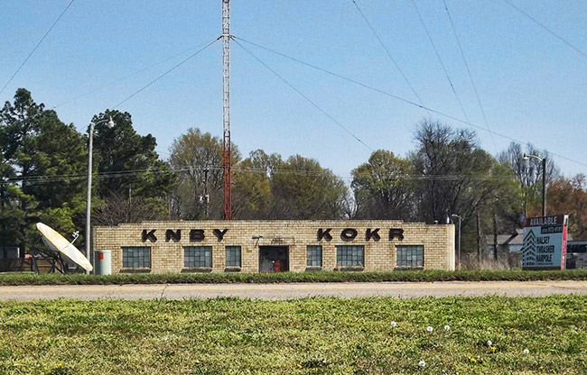 Single-story brick building with letters saying K.N.B.Y. and K.O.K.R. on it on street with radio tower behind it