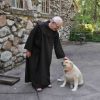 Older white man in religious robe and sandals petting a dog outside stone building