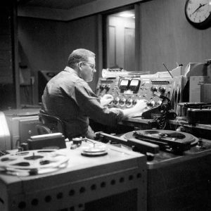 White man with glasses sitting at the control board in a radio station
