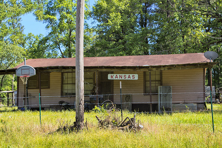 Single-story house inside barbed wire fence with hanging "Kansas" sign