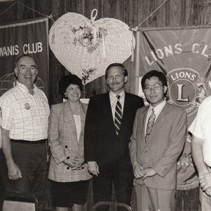 Two men in suits and woman in jacket with three men more casually dressed in front of banners