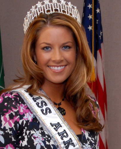 Young white woman with crown and "Miss Arkansas" sash smiling with American flag behind her
