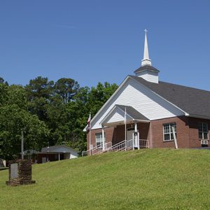 Brick church building with steeple and covered porch with parking lot and signs near the road in front of it