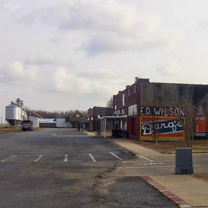 Brick storefronts silver grain silo and truck on parking lot and sign painted on the side of the building saying "Ed Wilson Barq's"