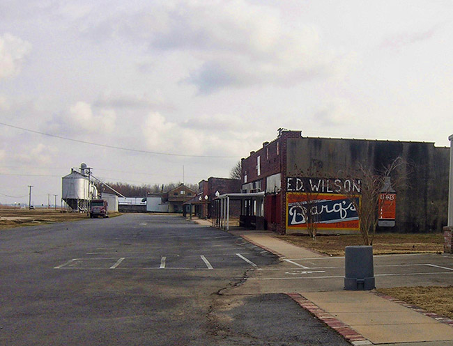 Brick storefronts silver grain silo and truck on parking lot and sign painted on the side of the building saying "Ed Wilson Barq's"