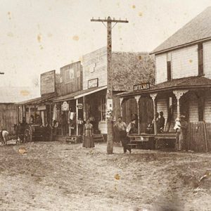 Old faded photo of storefronts with patrons and horses on dirt road with power lines