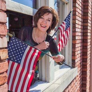 White woman smiling hanging out of window with American flags in both hands