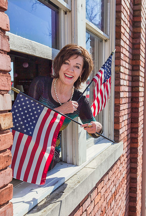 White woman smiling hanging out of window with American flags in both hands
