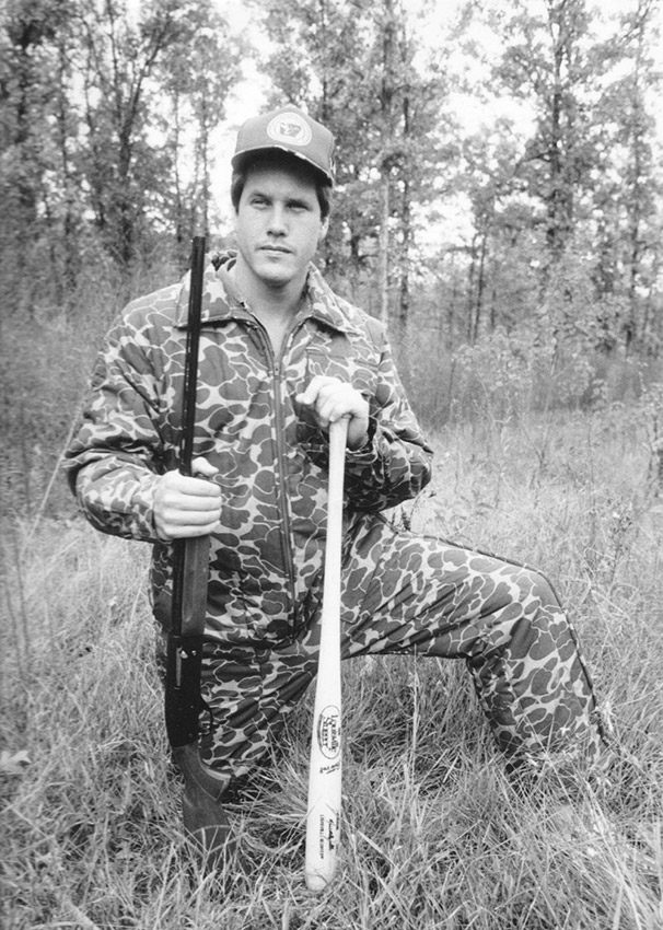 White man wearing camouflage and holding baseball bat and shotgun