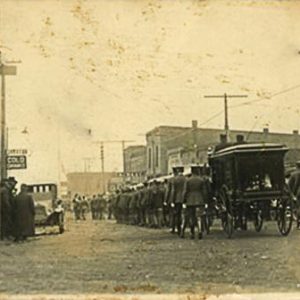 Line of people and funeral cart on dirt road with cars