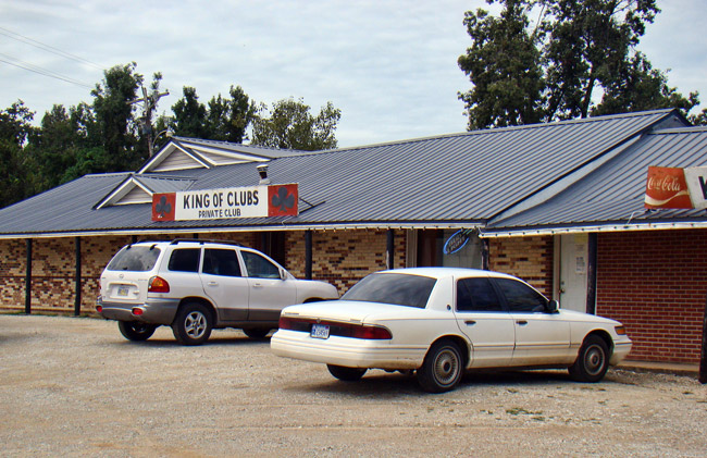 Single-story brick building with "King of Clubs private club" sign on gravel parking lot with parked cars