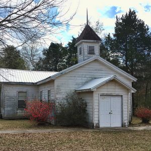 Weathered single-story church building with cupola surrounded by trees