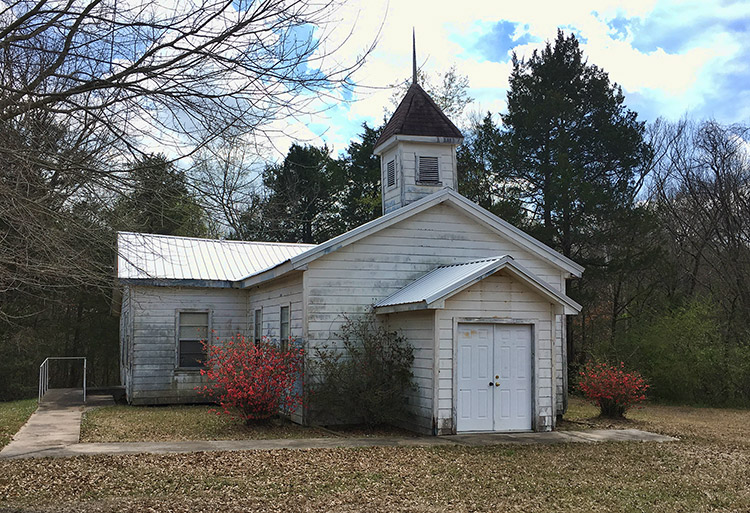 Weathered single-story church building with cupola surrounded by trees