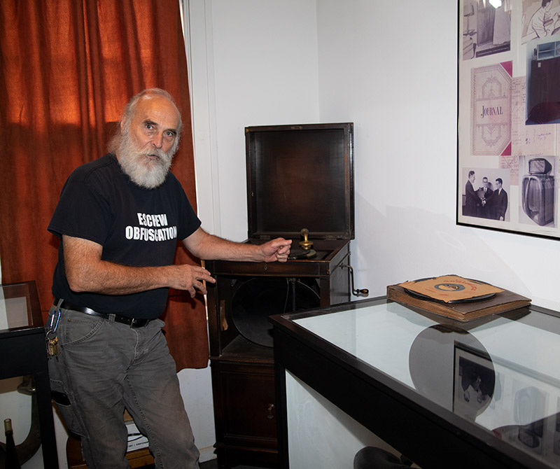 Older white man with beard and mustache in black shirt  and jeans standing next to phonograph player
